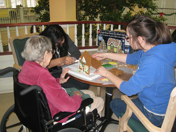 Students and a senior building a gingerbread house together Intergenerational volunteers building a gingerbread house